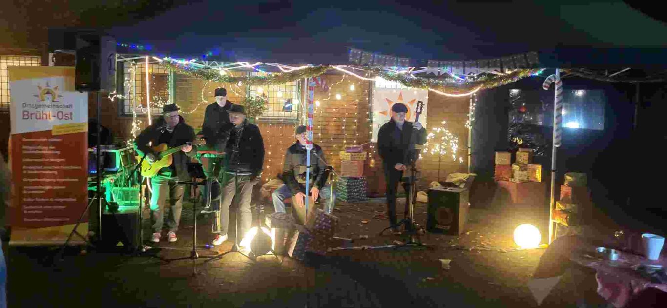 Weihnachtsmarkt Ortsgemeinschaft Brühl-Ost 2025 auf dem Vorplatz St. Stephan. Die Rhingpirate stimmen kölsche Weihnachtslieder an. Musikgruppe spielt live unter einem Zelt, umgeben von Lichtern und Dekoration.