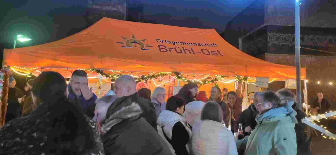 Weihnachtsmarkt Ortsgemeinschaft Brühl-Ost 2025 auf dem Vorplatz St. Stephan. Ausschank von Glühwein, Kakao und Kinderpunsch. Eine Menschenmenge versammelt sich unter einem orangefarbenen Festzelt bei Dunkelheit.