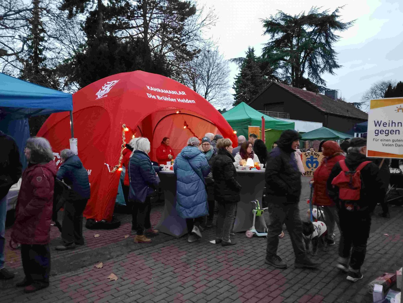 Weihnachtsmarkt Ortsgemeinschaft Brühl-Ost 2025 auf dem Vorplatz St. Stephan. Stand Kahramanlar Menschen stehen an Ständen unter bunten Zelten, während es dämmerig ist.
