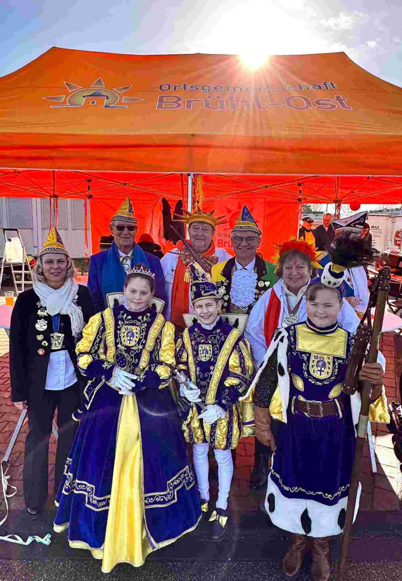 Das Kinderdreigestirn und der Bürgermeister Marc Prokop zu Besuch bei der Ortsgemeinschaft - Karnevalszug in Brühl-Ost 2026 Gruppe von Menschen in festlichen Kostümen vor einem orangen Zelt.