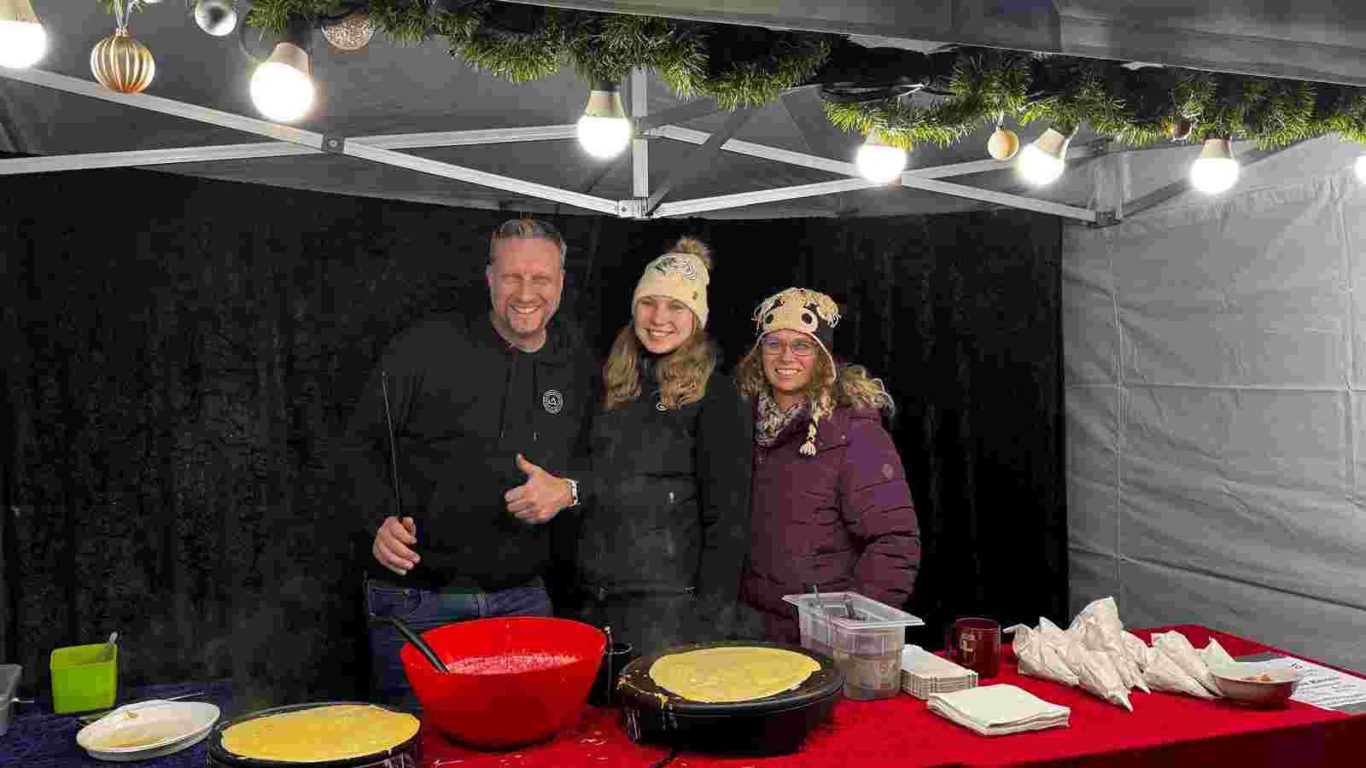 Der Modern Music School Stand auf dem Weihnachtsmarkt Ortsgemeinschaft Brühl-Ost 2025, Vorplatz St. Stephan Drei Personen stehen an einem Weihnachtsmarktstand mit einem großen roten Kochtopf.