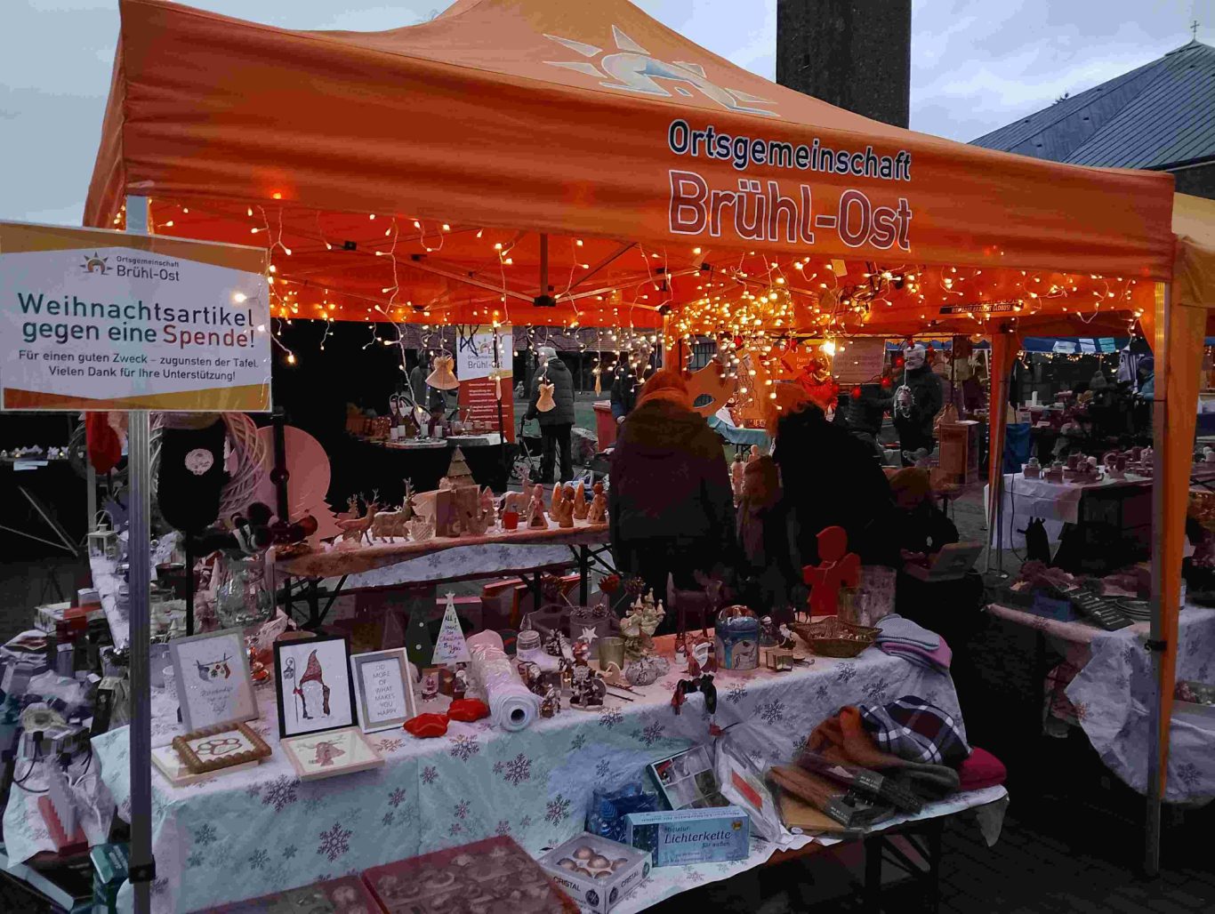 Spendenstand 2025 Weihnachtsmarkt Ortsgemeinschaft Brühl-Ost auf dem Vorplatz St. Stephan Bunte Verkaufsstände unter einem orangefarbenen Zelt mit Lichterketten und handgemachten Waren.