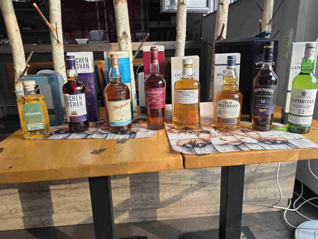 Ortsgemeinschaft Brühl-Ost. Einladung zum Whisky Tasting Verschiedene Flaschen Spirituosen auf einem Tisch, umgeben von hölzernen Dekorationen.